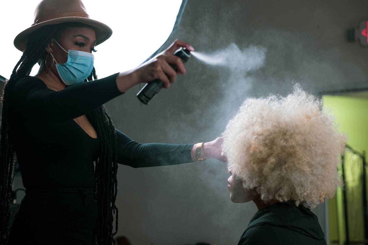 Stylist in mask sprays hair of seated client in studio setting.